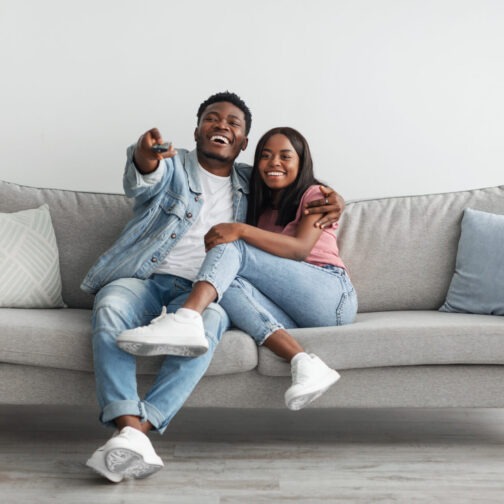 Beautiful African American black couple relaxing on sofa at home in living room, smiling man and woman spending free time together, watching TV or movie, embracing, laughing guy holding remote control